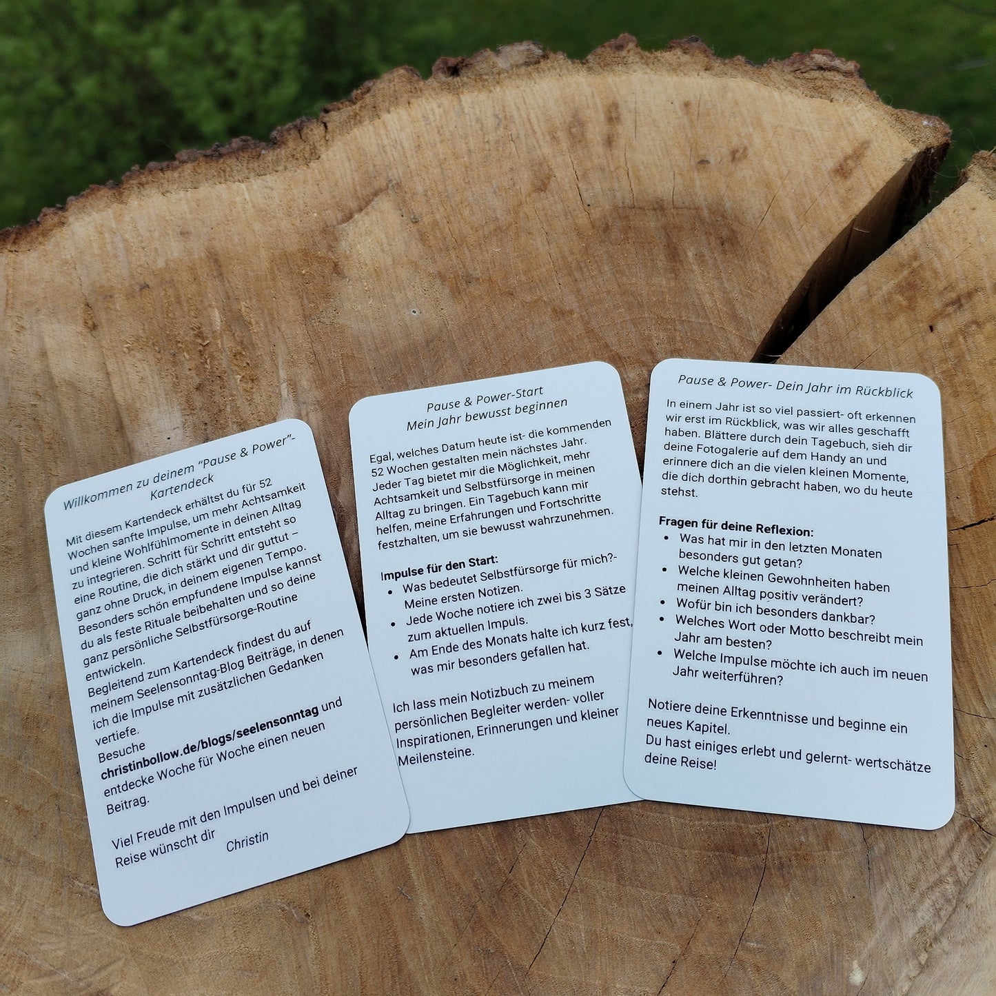 Kartendeck Pause und Power, Rückseite mit Text der Deckkarte, Reflektionskarte Start und Jahresrückblick. Die Karten liegen auf einer Holzscheibe im natürlichen Licht fotografiert, im Hintergrund Natur.