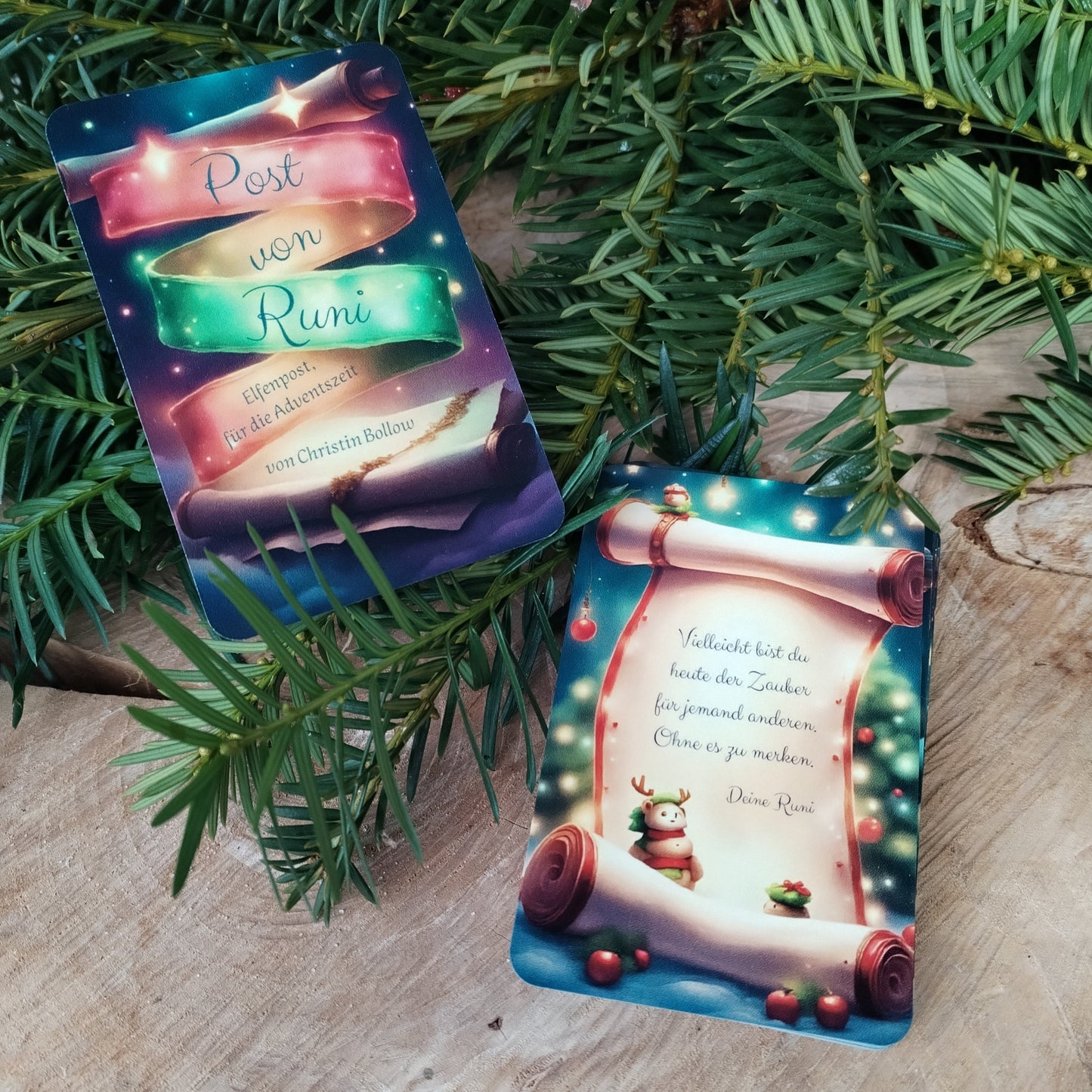 Kartendeck 'Post von Runi'. Das Dezemberdeck liegt mit Deckkarte und der Vorderseite der Impulskarten auf einer Holzscheibe und frischen Nadelbaumzweigen. Die Vorderseite der Karten zeigt ein winterliches Motiv mit einer aufgerollten Schriftrollen, auf der der Text von Winterelfe Runi steht, mit weiteren winterlich- weihnachtlichen Motiven im Hintergrund.
