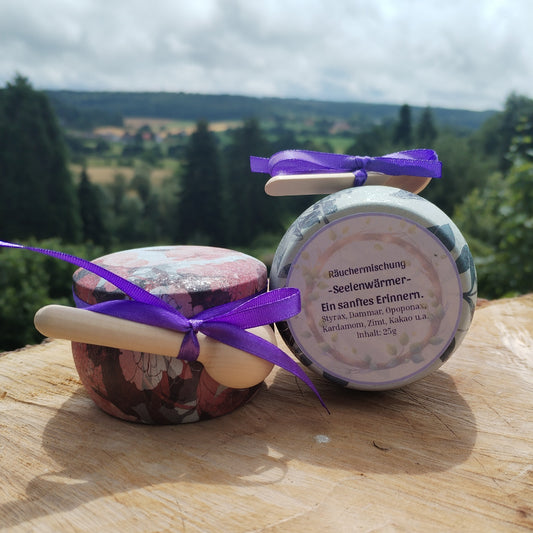 Produkt Räuchermischung Seelenwärmer, abgefüllt in einer bunten Metalldose, die auf einer Holzscheibe steht mit Natur im Hintergrund. Bild 2