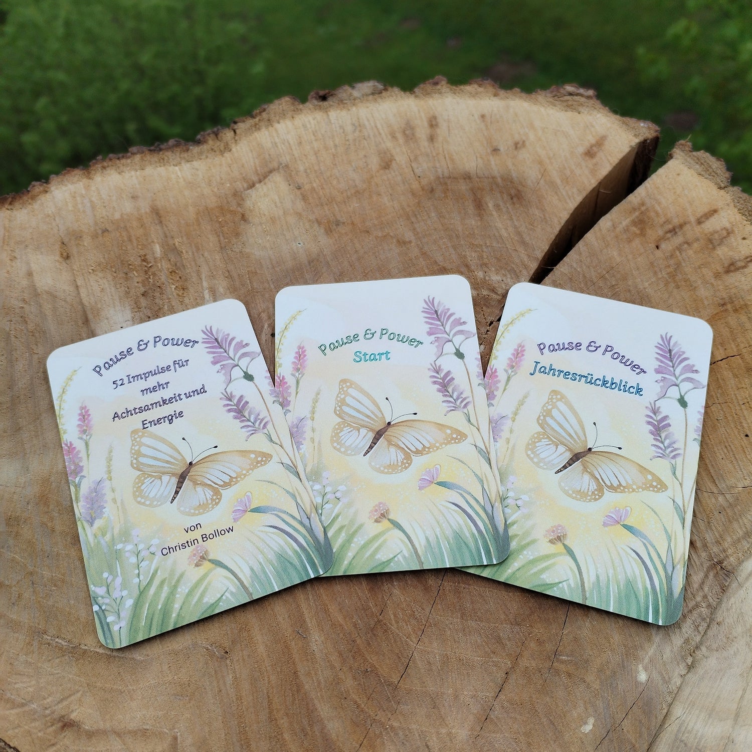 Kartendeck Pause und Power: Vorderseite mit Schmetterling und fliederfarbenen Blumen auf zartgelbem Hintergrund. Karten auf Holzscheibe im Naturlicht fotografiert.