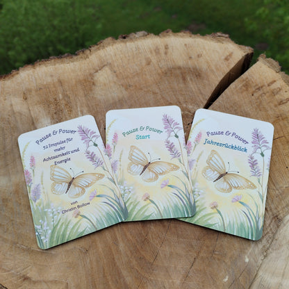 Kartendeck Pause und Power: Vorderseite mit Schmetterling und fliederfarbenen Blumen auf zartgelbem Hintergrund. Karten auf Holzscheibe im Naturlicht fotografiert.